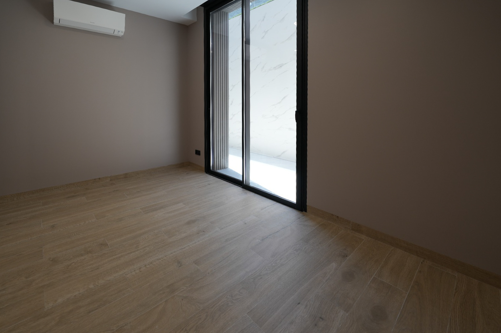 Pièce vide avec carrelage imitation bois clair, murs taupe et grande porte coulissante vitrée noire. Une unité de climatisation murale est présente. Pièce vide avec carrelage imitation bois clair, murs taupe et grande porte coulissante vitrée noire. Une unité de climatisation murale est présente.