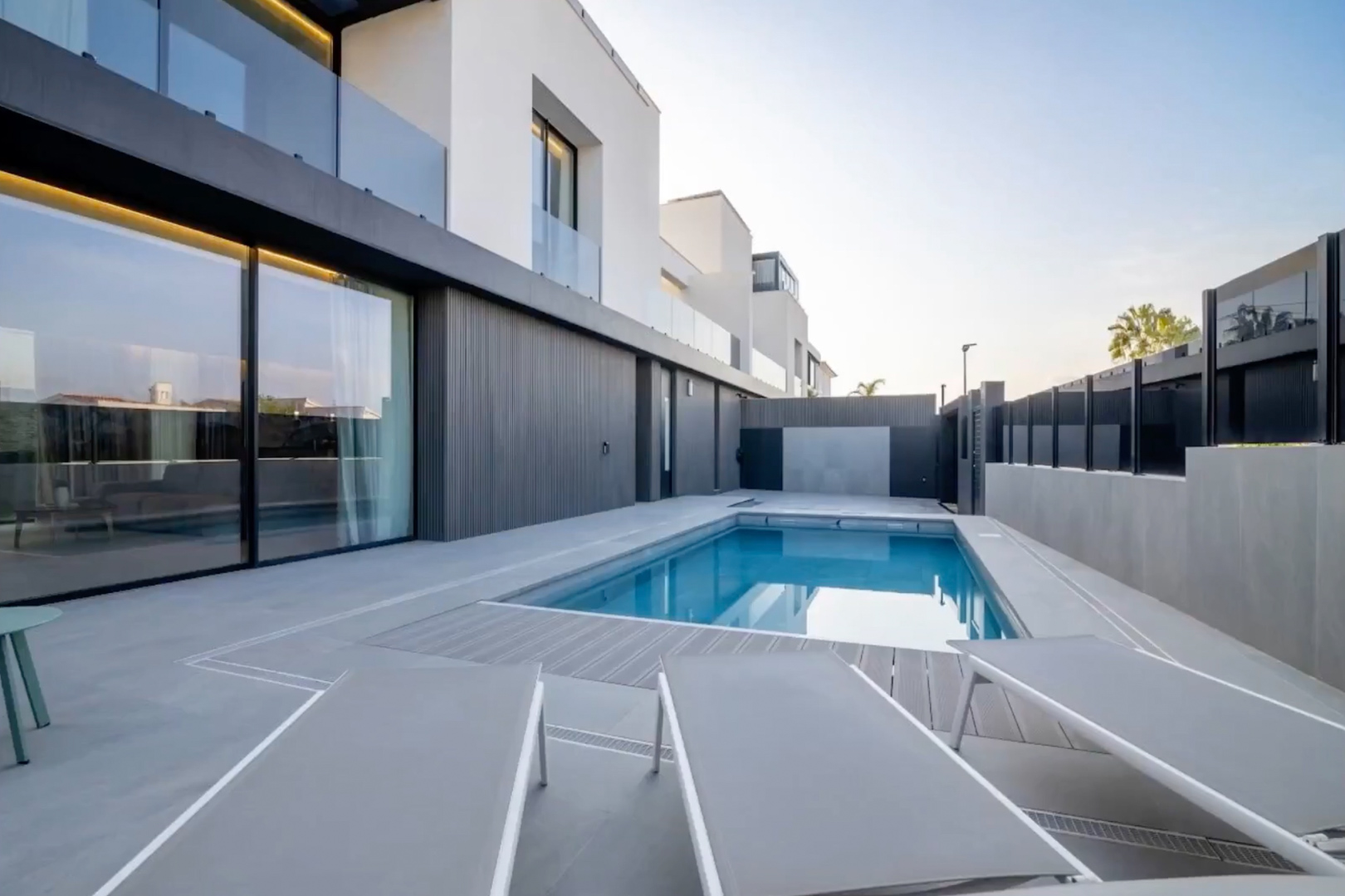 Piscine extérieure à l'eau bleue, entourée d'une terrasse carrelée grise et de platelage composite. Façade moderne avec de grandes baies vitrées reflétant le ciel. Piscine extérieure à l'eau bleue, entourée d'une terrasse carrelée grise et de platelage composite. Façade moderne avec de grandes baies vitrées reflétant le ciel.