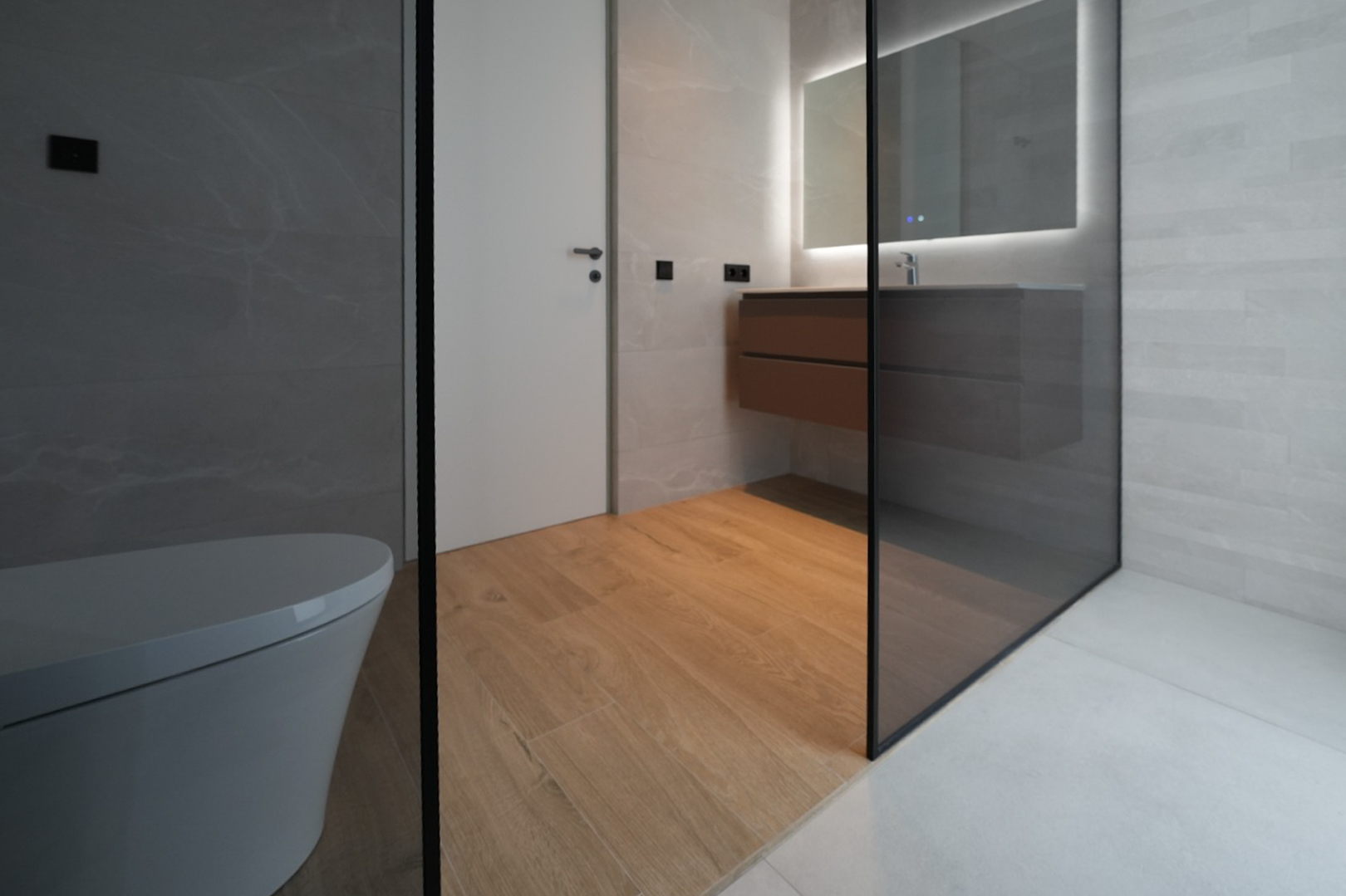 Salle de bain moderne avec toilettes suspendues, meuble vasque flottant en bois, miroir éclairé et douche vitrée. Le sol imitation bois contraste avec les carrelages muraux gris. Salle de bain moderne avec toilettes suspendues, meuble vasque flottant en bois, miroir éclairé et douche vitrée. Le sol imitation bois contraste avec les carrelages muraux gris.