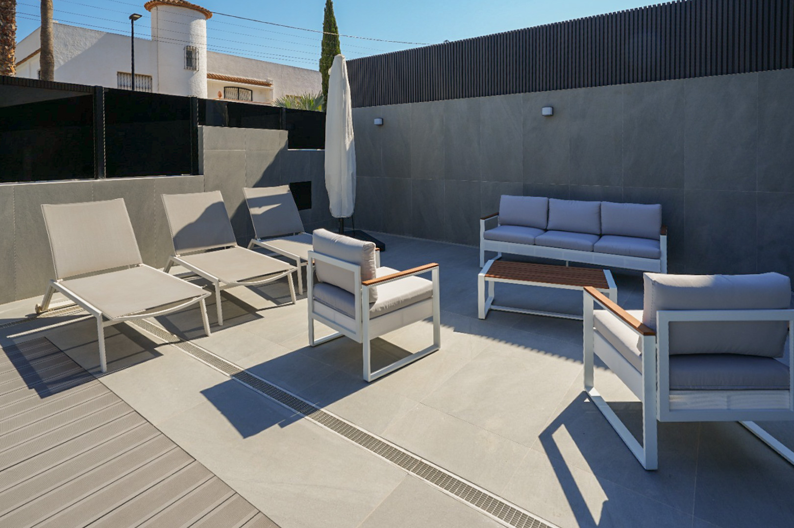 Terrasse extérieure aménagée avec mobilier contemporain blanc et gris, sol en carrelage et platelage composite. Comprend des transats et un salon de jardin. Terrasse extérieure aménagée avec mobilier contemporain blanc et gris, sol en carrelage et platelage composite. Comprend des transats et un salon de jardin.