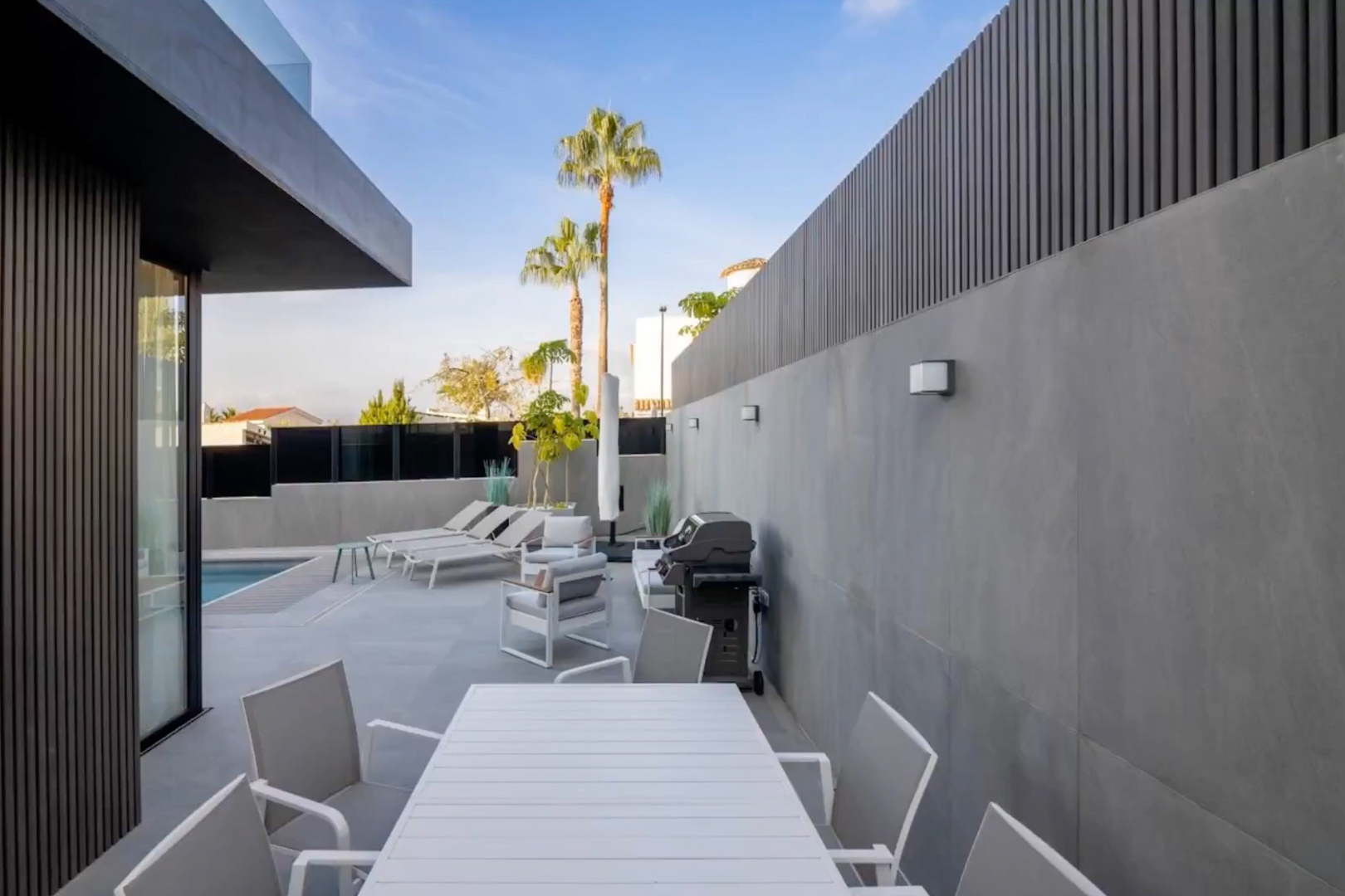 Terrasse extérieure moderne avec table à manger blanche à lattes, coin salon et barbecue, bordée de murs en carrelage gris et brise-vues sombres, avec vue sur la piscine. Terrasse extérieure moderne avec table à manger blanche à lattes, coin salon et barbecue, bordée de murs en carrelage gris et brise-vues sombres, avec vue sur la piscine.