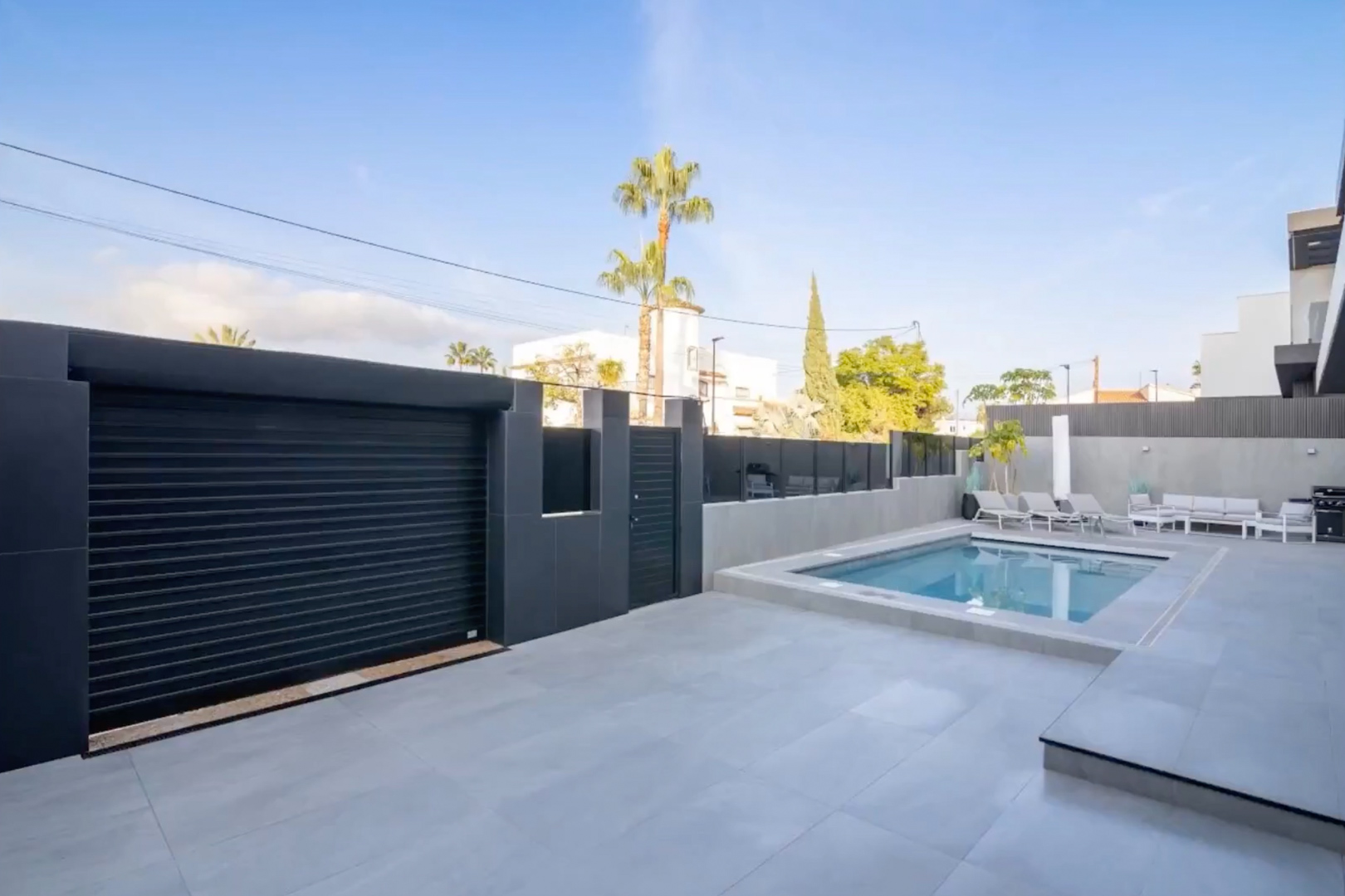 Vue extérieure de la piscine privée, du patio carrelé gris clair et du portail de garage sombre avec accès piéton sécurisé. Vue extérieure de la piscine privée, du patio carrelé gris clair et du portail de garage sombre avec accès piéton sécurisé.
