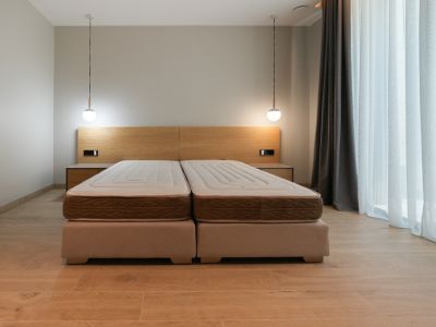 Chambre avec lit double composé de deux sommiers tapissiers, tête de lit en bois clair intégrant des tables de chevet et deux suspensions modernes. Grande fenêtre voilée.