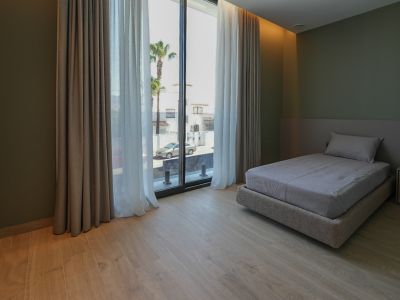 Chambre avec lit simple, parquet clair et murs vert olive. Les grandes baies vitrées avec doubles rideaux offrent une vue sur la rue et les palmiers.