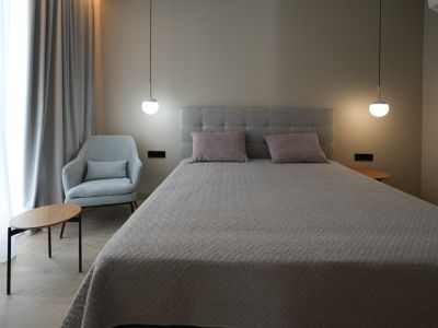 Chambre élégante avec un lit capitonné gris, deux suspensions modernes et un fauteuil confortable. Les rideaux clairs et le sol effet bois créent une ambiance sereine.