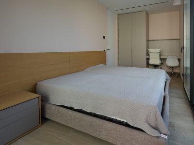 Chambre moderne avec lit capitonné gris clair, tête de lit en bois et table de chevet intégrée. Elle comprend une armoire encastrée, un double espace bureau et de grandes baies vitrées.