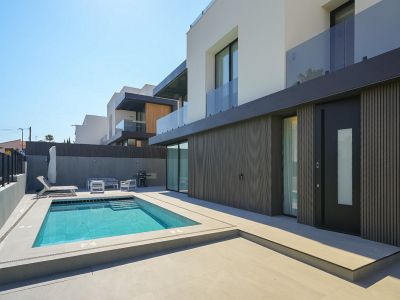Extérieur de villa moderne avec piscine rectangulaire et escaliers intégrés, entourée d'un dallage clair. Façade du rez-de-chaussée avec revêtement à lattes sombres et porte d'entrée distinctive.