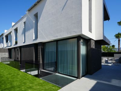 Façade moderne avec textures contrastées blanc/gris et sombre, grandes baies vitrées, pelouse artificielle et terrasse carrelée aménagée avec mobilier extérieur.