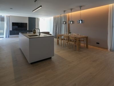 Îlot de cuisine moderne avec évier intégré et plaque de cuisson, ouvert sur une salle à manger avec table en bois et luminaires suspendus. Sol carrelé imitation bois clair.