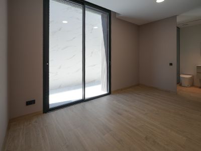 Pièce spacieuse avec sol carrelé imitation bois et murs clairs. De grandes portes coulissantes vitrées noires mènent à une terrasse extérieure carrelée de marbre blanc. Salle de bain intégrée visible.
