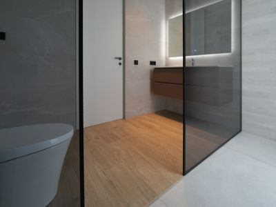 Salle de bain moderne avec toilettes suspendues, meuble vasque flottant en bois, miroir éclairé et douche vitrée. Le sol imitation bois contraste avec les carrelages muraux gris.