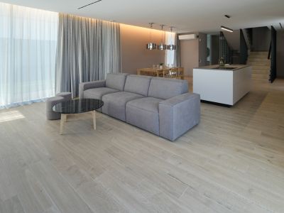 Salon ouvert avec canapé modulaire gris, table basse en marbre noir et sol carrelé aspect bois. Grandes fenêtres avec voilages et rideaux gris.