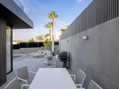 Terrasse extérieure moderne avec table à manger blanche à lattes, coin salon et barbecue, bordée de murs en carrelage gris et brise-vues sombres, avec vue sur la piscine.
