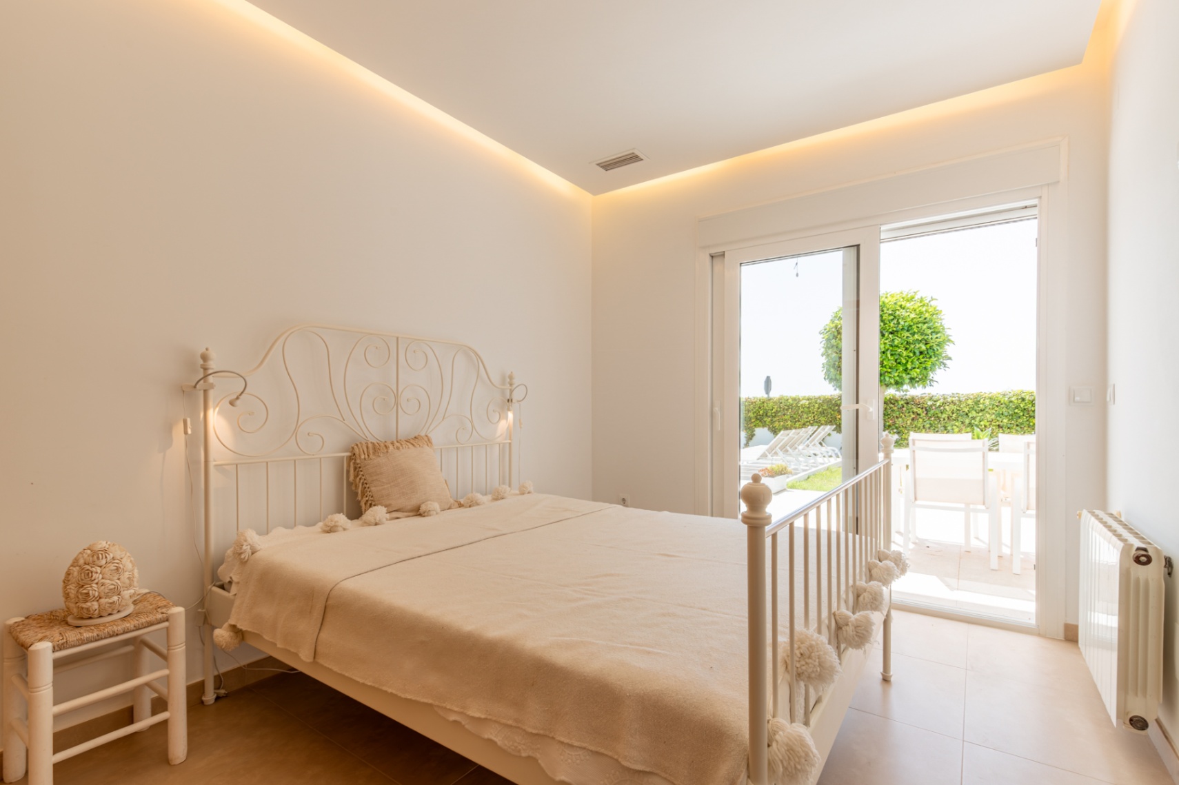 Chambre lumineuse avec lit en fer forgé blanc, éclairage LED indirect et accès direct à une terrasse privée avec jardin.