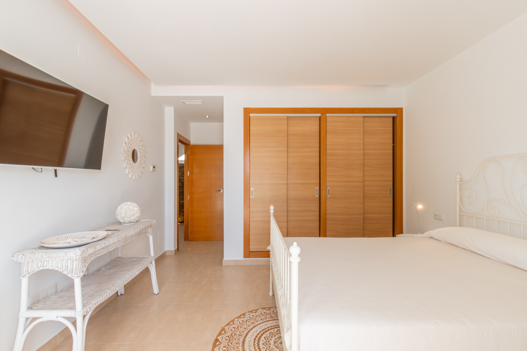 Chambre lumineuse avec lit en fer forgé blanc, placards intégrés en bois clair et télévision murale. L'espace comprend une console en osier et un sol carrelé.