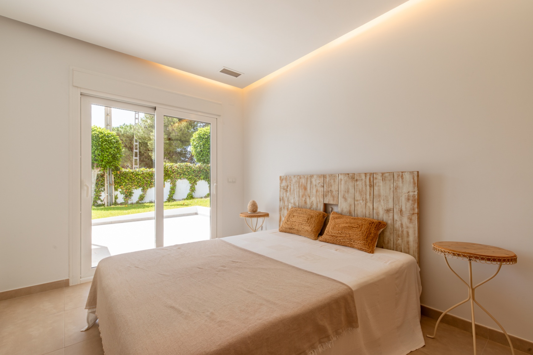 Chambre lumineuse avec tête de lit en bois rustique, éclairage intégré au plafond et accès direct à une terrasse jardin privée par des portes coulissantes.