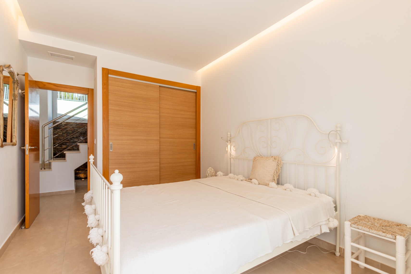 Chambre lumineuse avec un lit en fer forgé blanc, une armoire intégrée en bois clair et un accès à l'escalier intérieur. Éclairage LED au plafond.