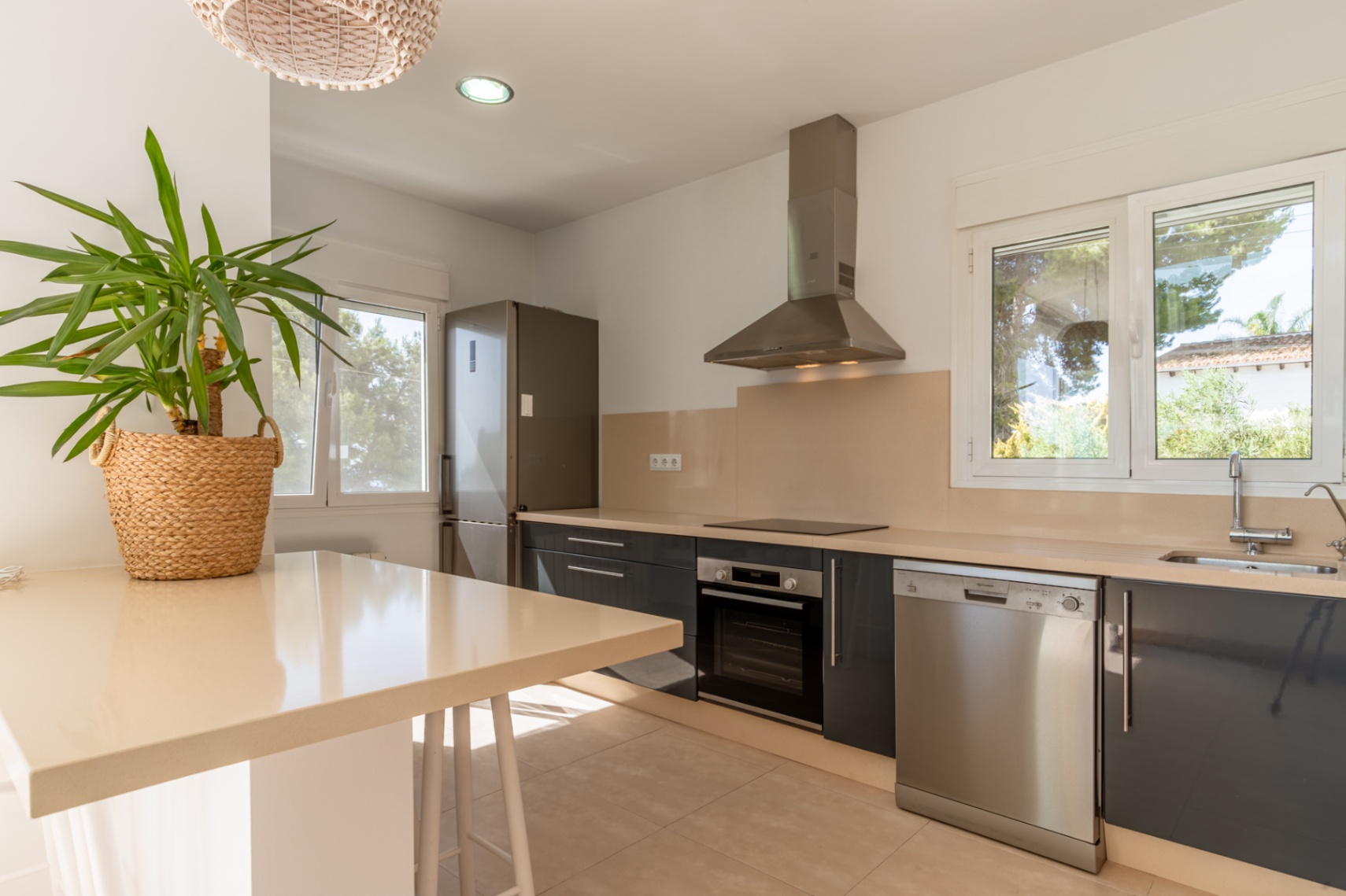 Cuisine moderne avec des armoires gris foncé, des plans de travail beige clair et des appareils en acier inoxydable. De grandes fenêtres offrent une lumière naturelle.