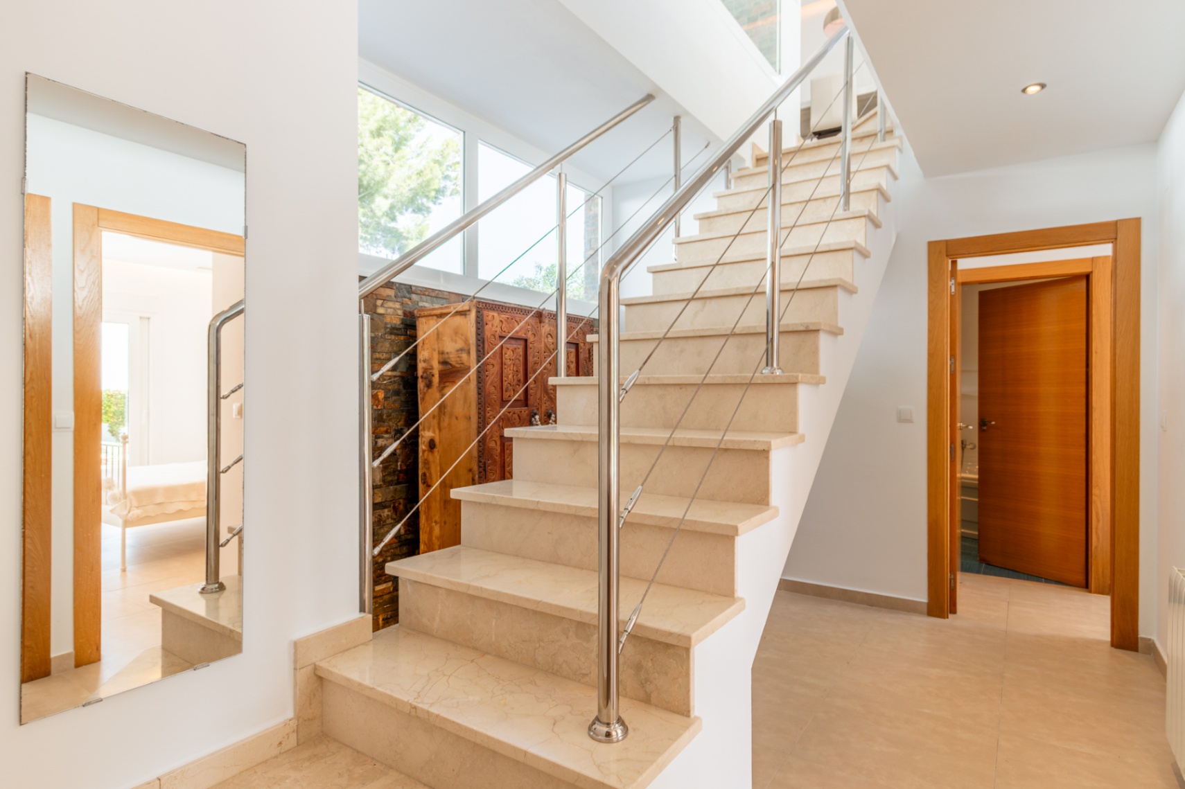 Escalier intérieur en marbre clair avec rampe en acier inoxydable. Les grandes fenêtres éclairent un meuble en bois sculpté contre un mur en pierre, un miroir reflète une pièce adjacente.