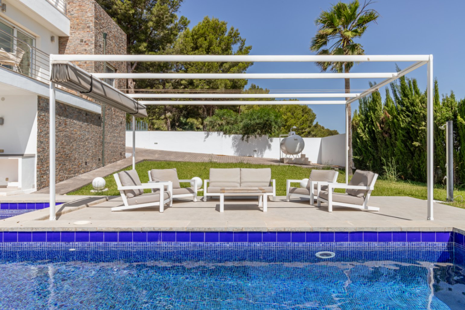 Espace lounge extérieur moderne sous pergola blanche avec store rétractable, sur une terrasse pavée à côté de la piscine en mosaïque bleue et des jardins.