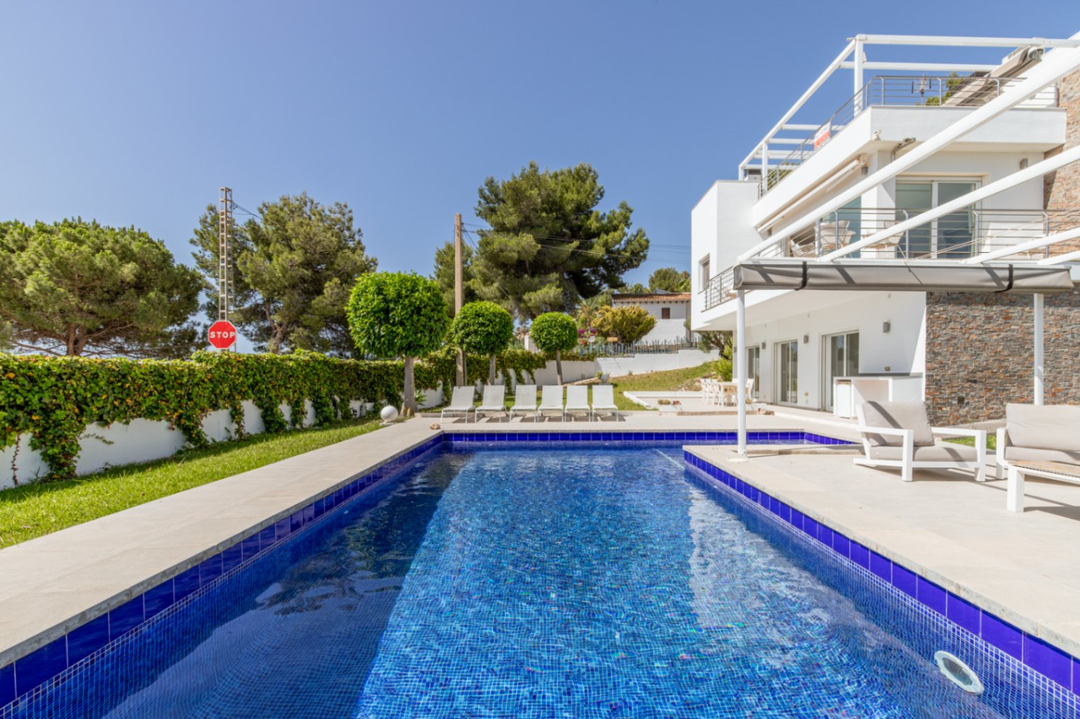 La piscine de 9x4 mètres aux carreaux de mosaïque bleus est entourée d'une terrasse claire avec chaises longues, jouxtant la villa blanche moderne avec parement en pierre et terrasse ombragée.