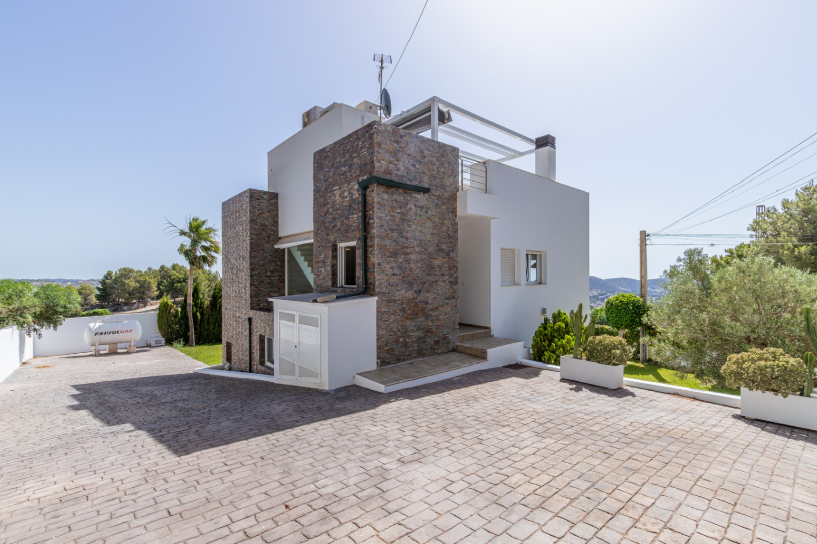 L'extérieur moderne de la villa combine parement en pierre, une large allée pavée et une rampe d'accès. Un réservoir de gaz Repsol est visible, avec un aménagement paysager méditerranéen.