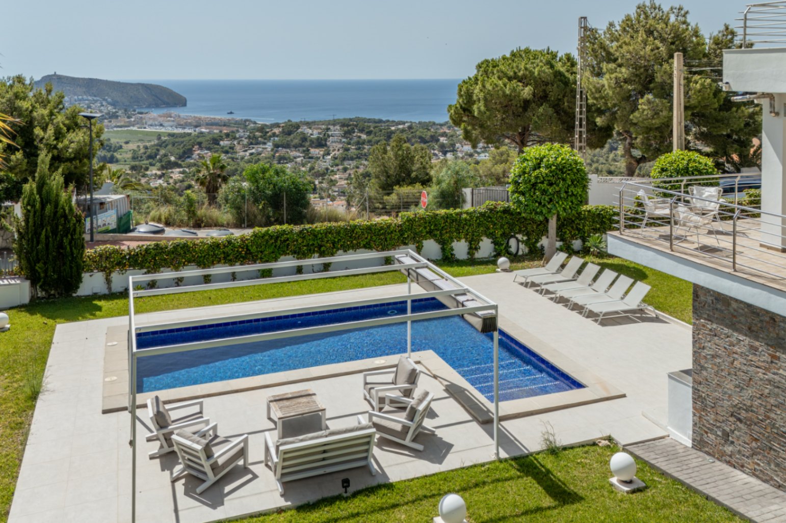 Piscine avec couverture automatique et salon extérieur sous pergola, entourée de jardins soignés avec vue panoramique sur la mer de Moraira.