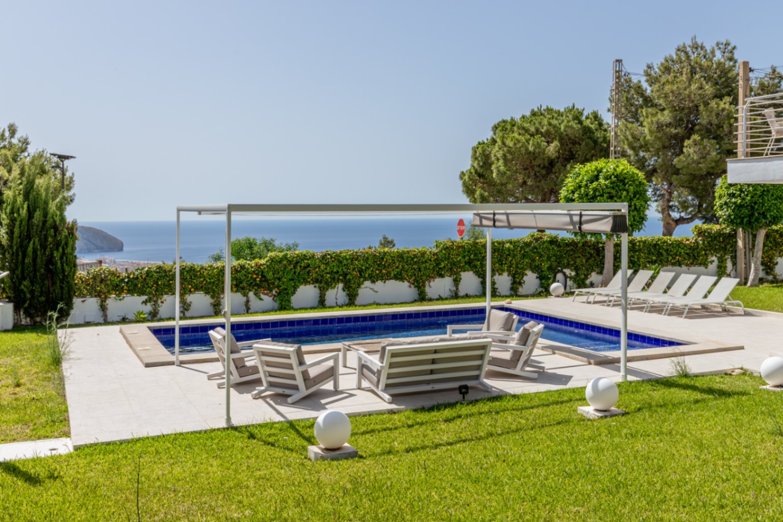 Piscine carrelée de bleu et terrasse pavée avec mobilier de salon sous une pergola blanche. Pelouses soignées encadrant les vues dégagées sur la mer.