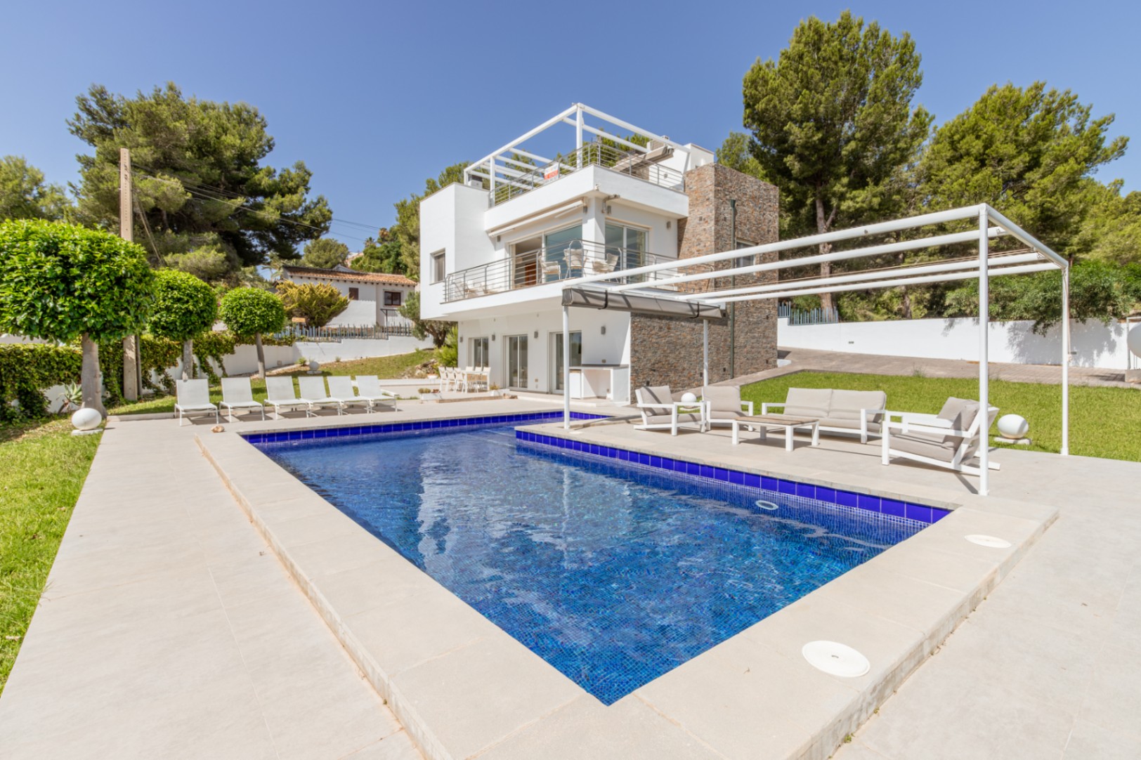 Piscine en mosaïque bleue et terrasse pavée avec espace détente sous pergola, devant la villa moderne aux façades blanches et pierre.