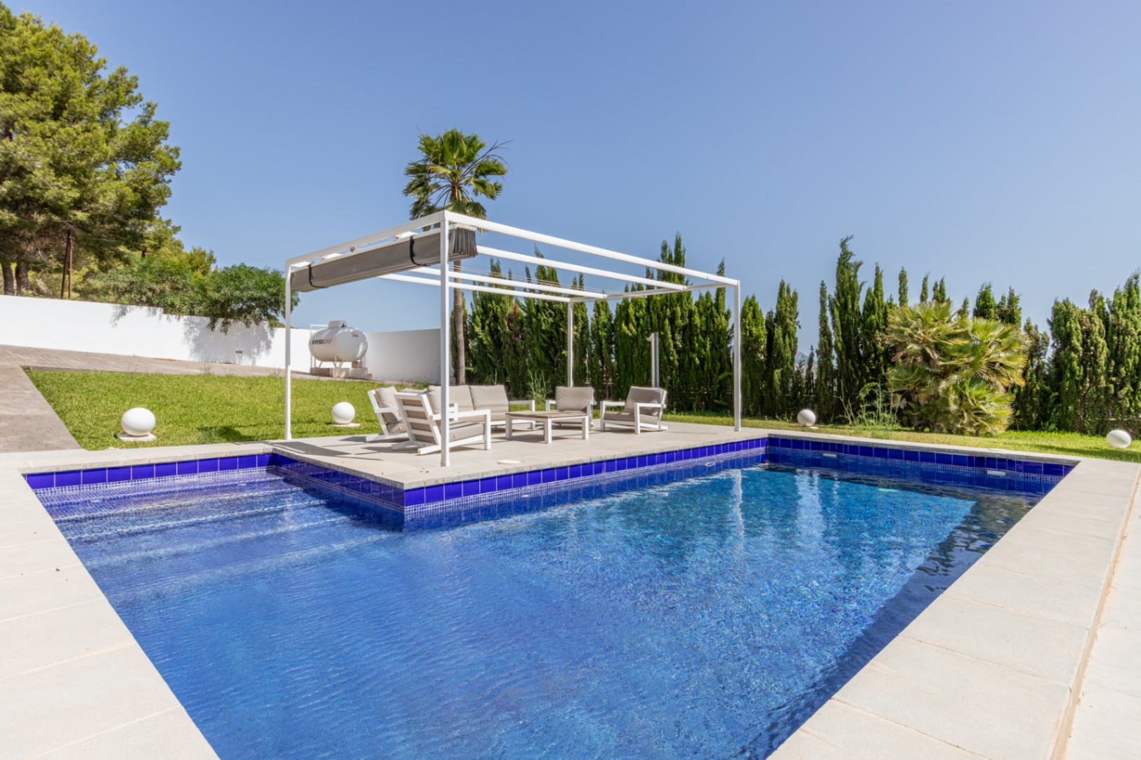 Piscine rectangulaire en mosaïque bleue avec escaliers, jouxtant une pergola blanche moderne et un salon de jardin sur pelouse entretenue.