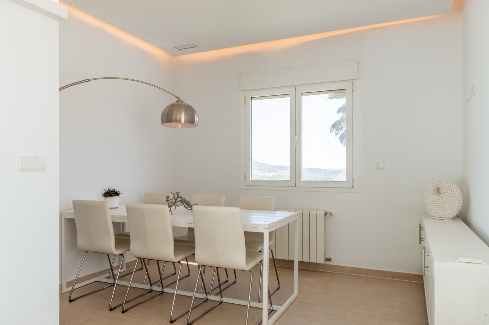 Salle à manger lumineuse avec table blanche pour six, lampadaire arqué et éclairage d'ambiance au plafond. Les fenêtres offrent des vues sur la verdure et les collines.