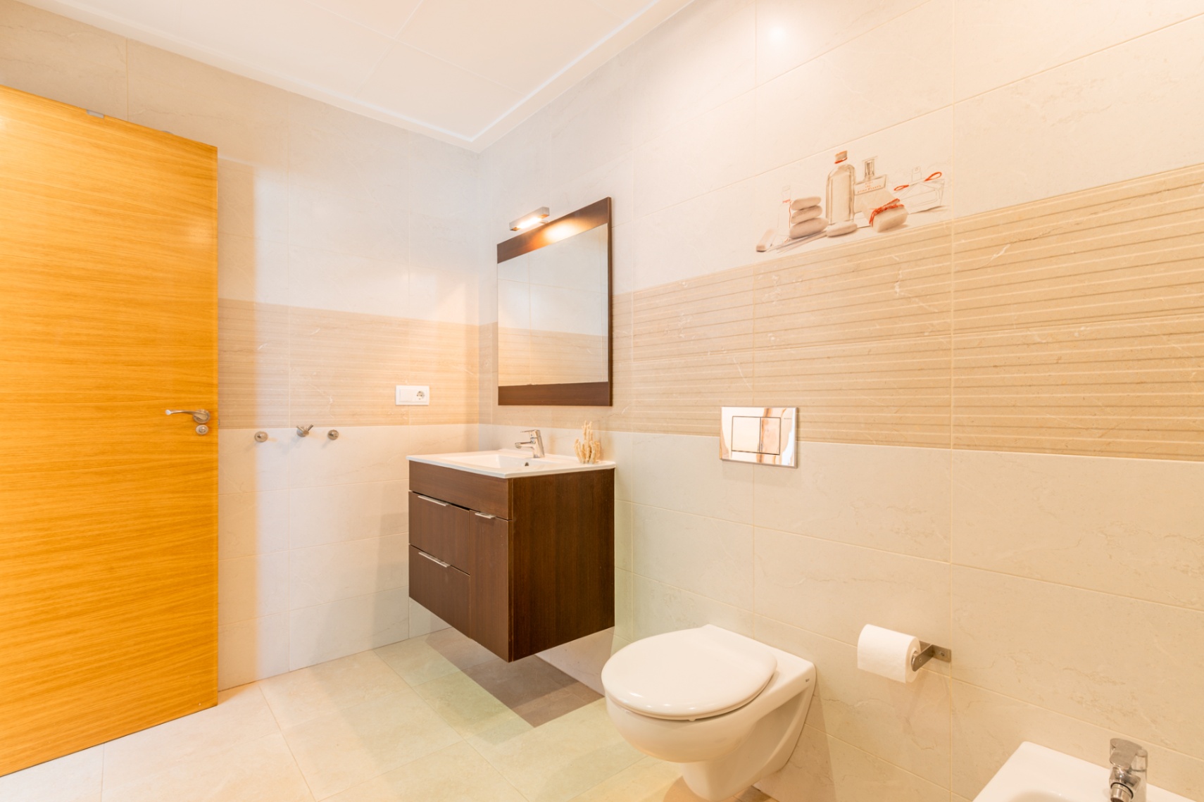 Salle de bain contemporaine avec carrelage clair, porte en bois, meuble-lavabo suspendu en bois foncé et miroir. WC mural avec plaque de chasse chromée.
