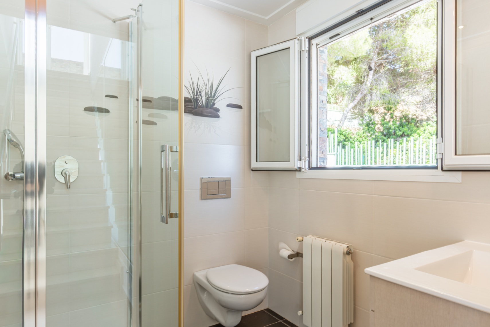 Salle de bain fonctionnelle avec douche vitrée, WC suspendu et fenêtre en aluminium blanc offrant une vue sur la verdure. Murs carrelés beiges et radiateur.