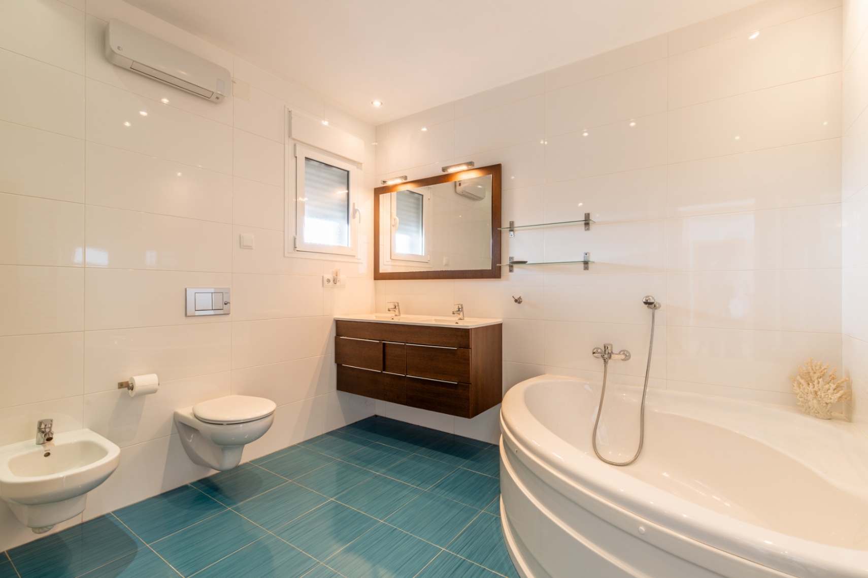 Salle de bain lumineuse avec baignoire d'angle, double vasque en bois foncé et WC suspendu. Carrelage blanc contrastant avec un sol turquoise vif, lumière naturelle et climatisation.
