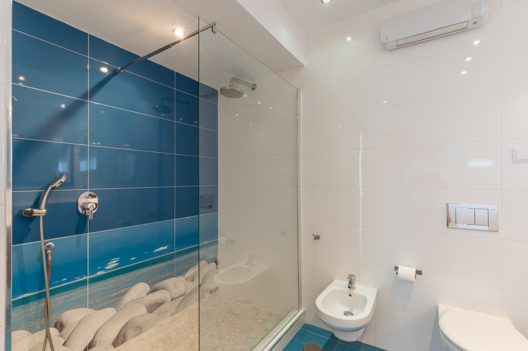 Salle de bain moderne avec douche à l'italienne et paroi vitrée, mur carrelé bleu avec motif plage de galets et sol en pierre. Comprend bidet et WC suspendu.