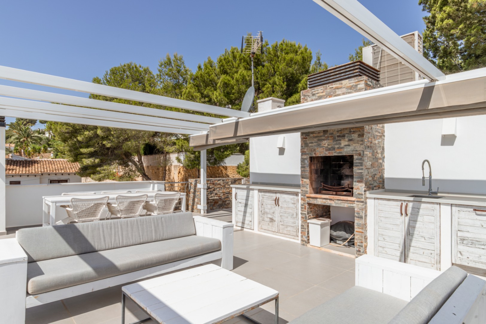 Solarium sur le toit avec cuisine d'été en pierre, barbecue intégré et évier, ainsi que des espaces détente et repas sous une pergola blanche, entouré d'arbres luxuriants.