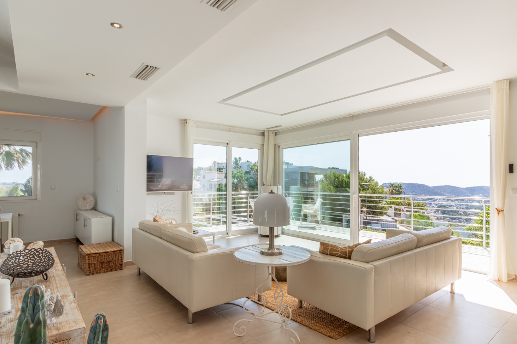 Vaste salon lumineux avec canapés clairs et grandes baies vitrées coulissantes donnant sur un balcon. Profitez de vues panoramiques sur les collines depuis cet espace doté d'une TV murale.