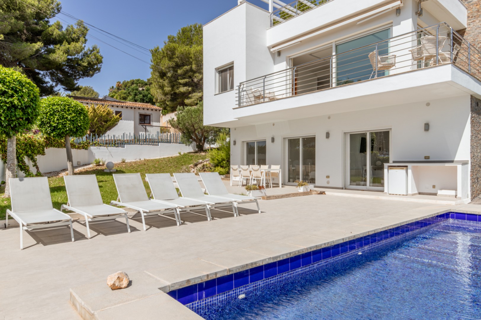 Vaste terrasse carrelée avec grande piscine en mosaïque bleue, six chaises longues blanches et coin repas extérieur. Une cuisine d'été intégrée avec réfrigérateur complète cet espace de loisirs.
