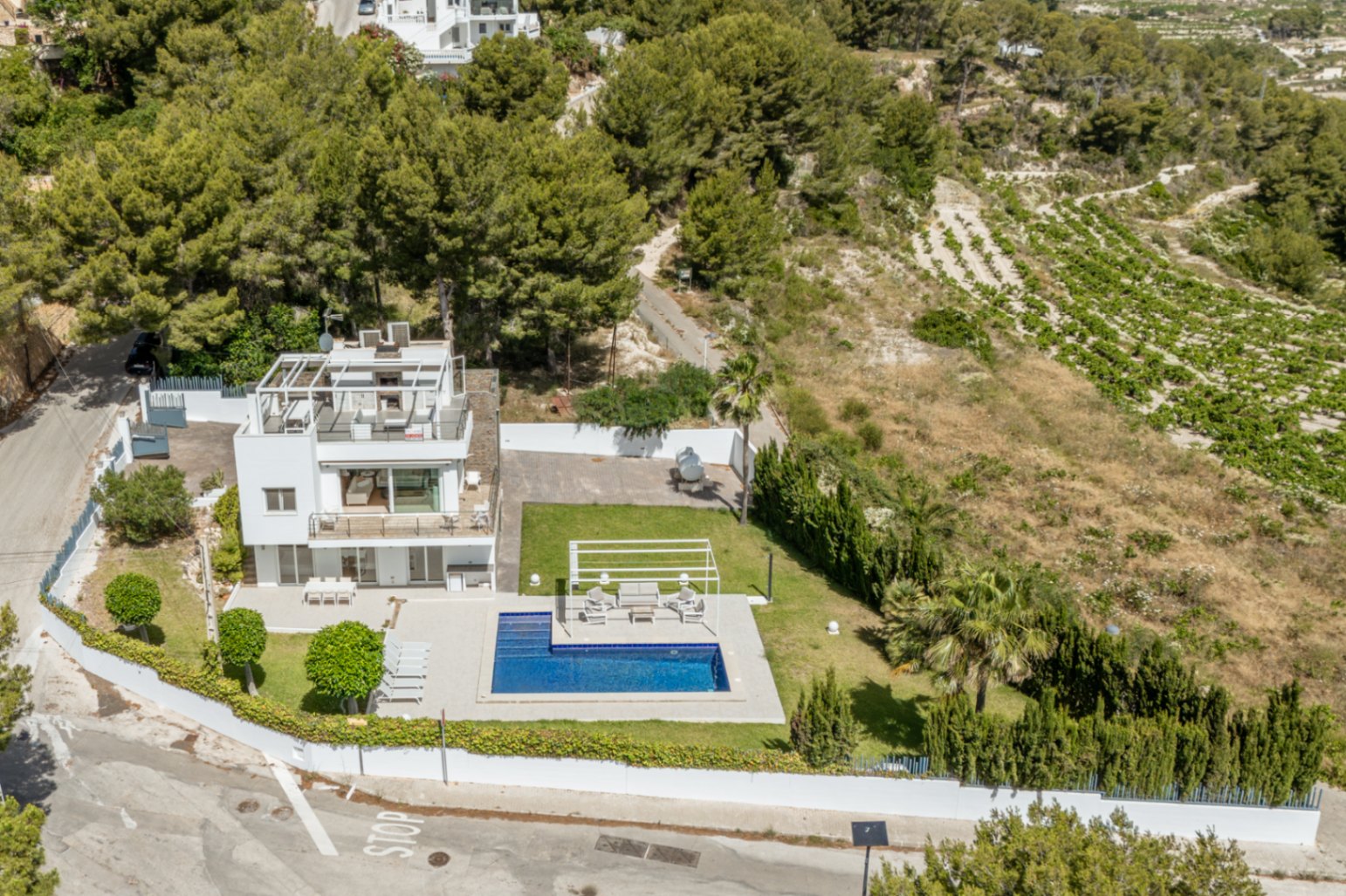 Vue aérienne de la villa blanche à trois niveaux, avec piscine, pelouse soignée et solarium sur le toit avec pergola. Entourée de pins et de vignobles.