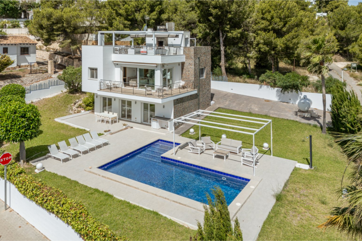 Vue aérienne de la villa moderne à plusieurs niveaux, avec piscine rectangulaire, vastes terrasses salon et salle à manger, et pelouses soignées, entourée de végétation luxuriante.