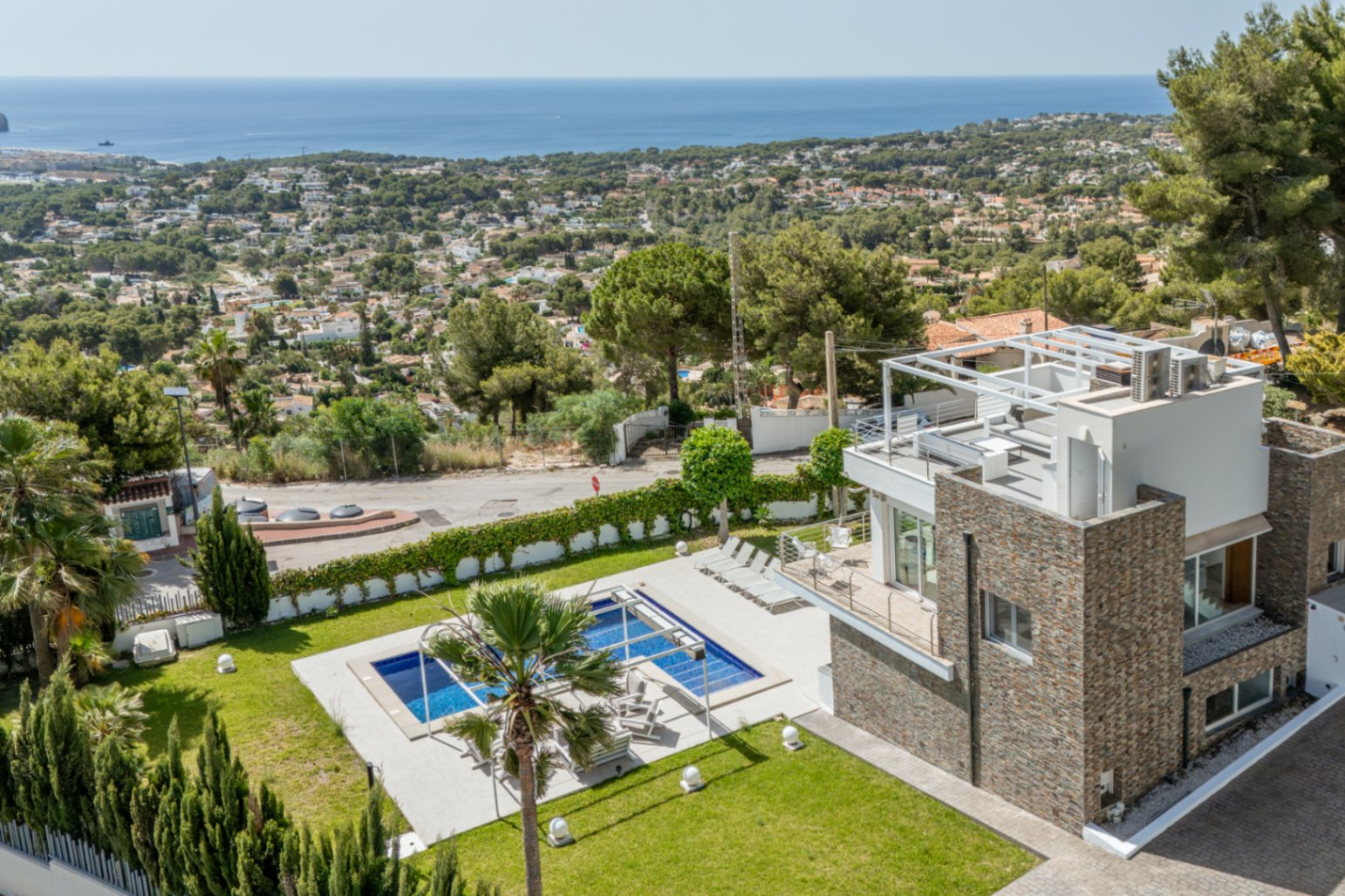 Vue aérienne de la villa moderne avec revêtement en pierre, piscine rectangulaire, pelouse soignée et solarium sur le toit avec pergola, offrant des vues mer panoramiques.