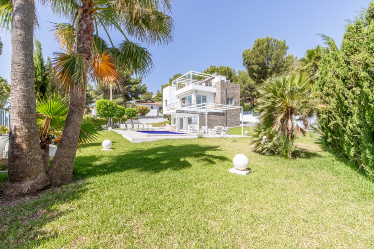 Vue extérieure de la villa moderne à plusieurs niveaux, avec grande piscine, pelouse impeccable, palmiers et luminaires sphériques.