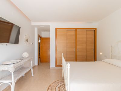 Chambre lumineuse avec lit en fer forgé blanc, placards intégrés en bois clair et télévision murale. L'espace comprend une console en osier et un sol carrelé.