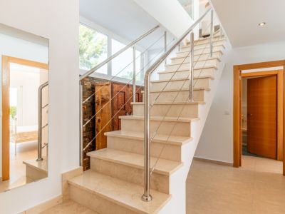 Escalier intérieur en marbre clair avec rampe en acier inoxydable. Les grandes fenêtres éclairent un meuble en bois sculpté contre un mur en pierre, un miroir reflète une pièce adjacente.