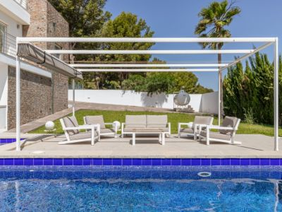 Espace lounge extérieur moderne sous pergola blanche avec store rétractable, sur une terrasse pavée à côté de la piscine en mosaïque bleue et des jardins.