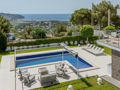 Piscine avec couverture automatique et salon extérieur sous pergola, entourée de jardins soignés avec vue panoramique sur la mer de Moraira.