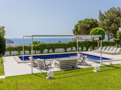 Piscine carrelée de bleu et terrasse pavée avec mobilier de salon sous une pergola blanche. Pelouses soignées encadrant les vues dégagées sur la mer.