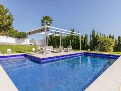 Piscine rectangulaire en mosaïque bleue avec escaliers, jouxtant une pergola blanche moderne et un salon de jardin sur pelouse entretenue.