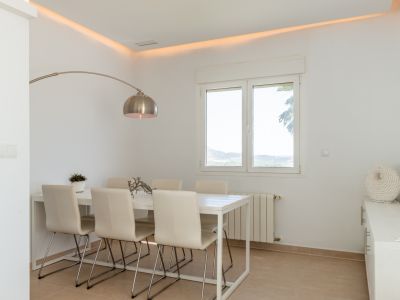 Salle à manger lumineuse avec table blanche pour six, lampadaire arqué et éclairage d'ambiance au plafond. Les fenêtres offrent des vues sur la verdure et les collines.