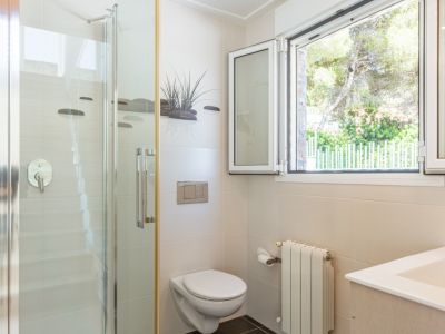 Salle de bain fonctionnelle avec douche vitrée, WC suspendu et fenêtre en aluminium blanc offrant une vue sur la verdure. Murs carrelés beiges et radiateur.